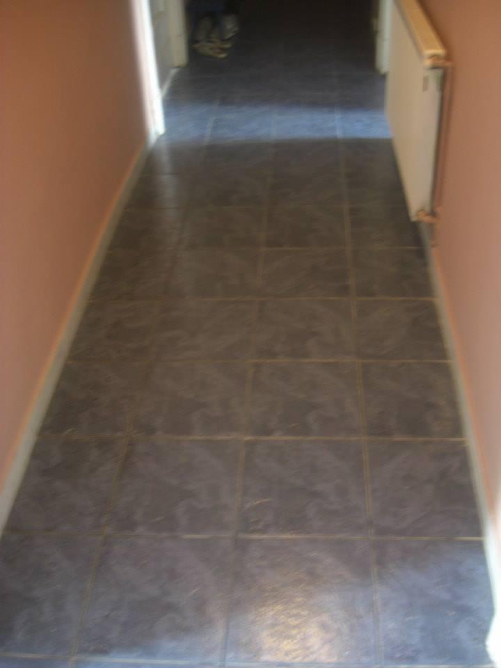 Widok korytarza z ciemnymi, kwadratowymi płytkami podłogowymi, różowe ściany i biały grzejnik po prawej stronie. W głębi korytarza widoczne buty.