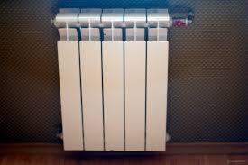 Biały, aluminiowy grzejnik panelowy zamontowany na ścianie z tapetą o drobnej, geometrycznej fakturze, widoczny fragment drewnianej podłogi.