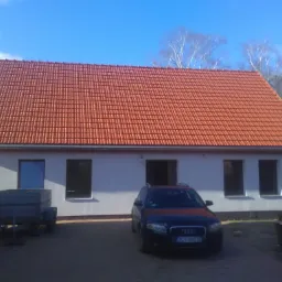 Nowo wybudowany dom z czerwoną dachówką, widoczny fragment przyczepy i samochód osobowy na podjeździe.