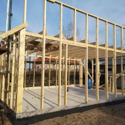 Wznoszenie konstrukcji szkieletowej domu z widocznym fundamentem, izolacją przeciwwilgociową i niebieską beczką w tle.