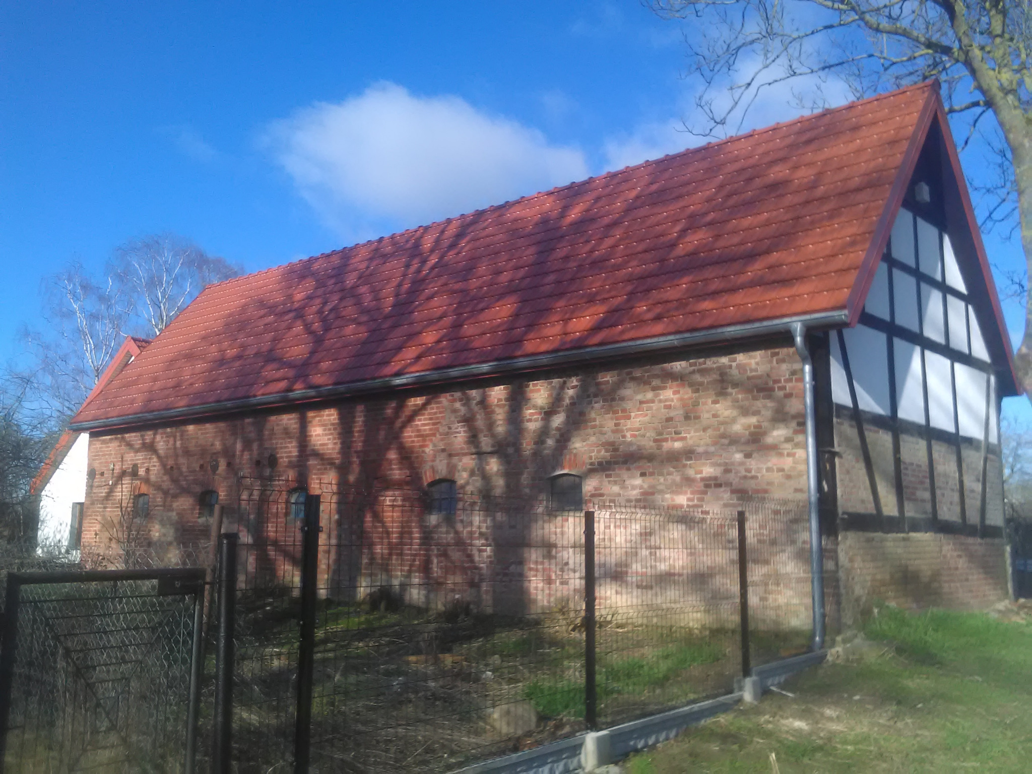 Budynek gospodarczy z czerwonym dachem krytym dachówką, częściowo murowany z cegły, częściowo konstrukcja szachulcowa, z widocznymi rynnami i cieniem drzew na elewacji