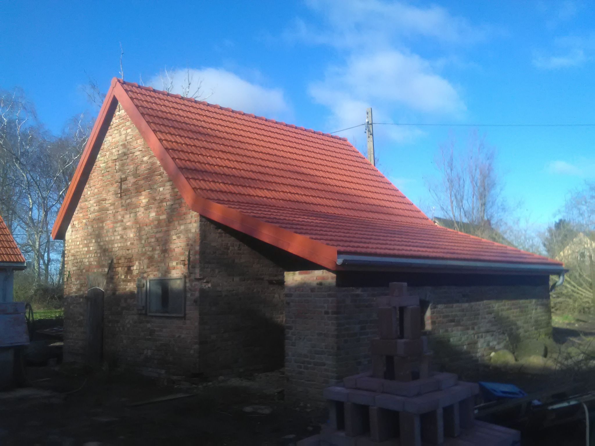 Budynek gospodarczy z czerwonej cegły z nowym, czerwonym dachem z dachówki ceramicznej, widok z zewnątrz w słoneczny dzień, na pierwszym planie ułożone kostki brukowe.