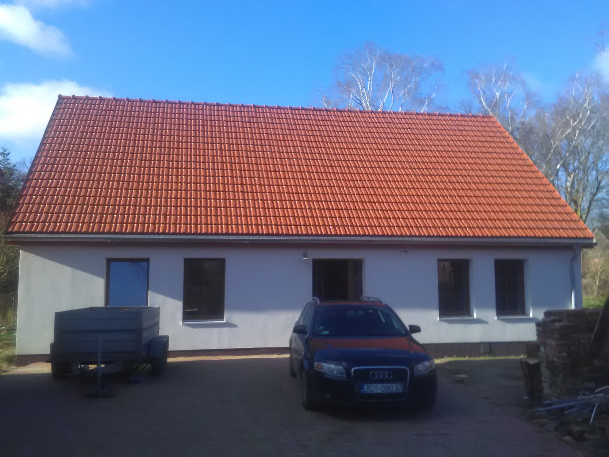 Nowo wybudowany dom z czerwoną dachówką, widoczny fragment przyczepy i samochód osobowy na podjeździe.