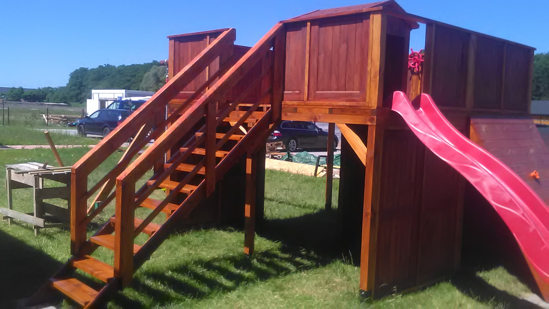 Drewniany plac zabaw z wieżą, schodami i czerwoną zjeżdżalnią na trawniku, widoczne samochody w tle.