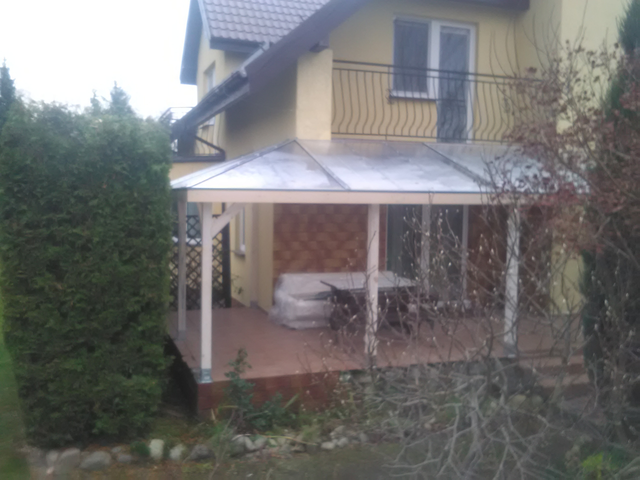 Zadaszony taras przy żółtym domu z balkonem, częściowo zasłonięty przez krzewy; widoczny stół i krzesła owinięte folią ochronną.