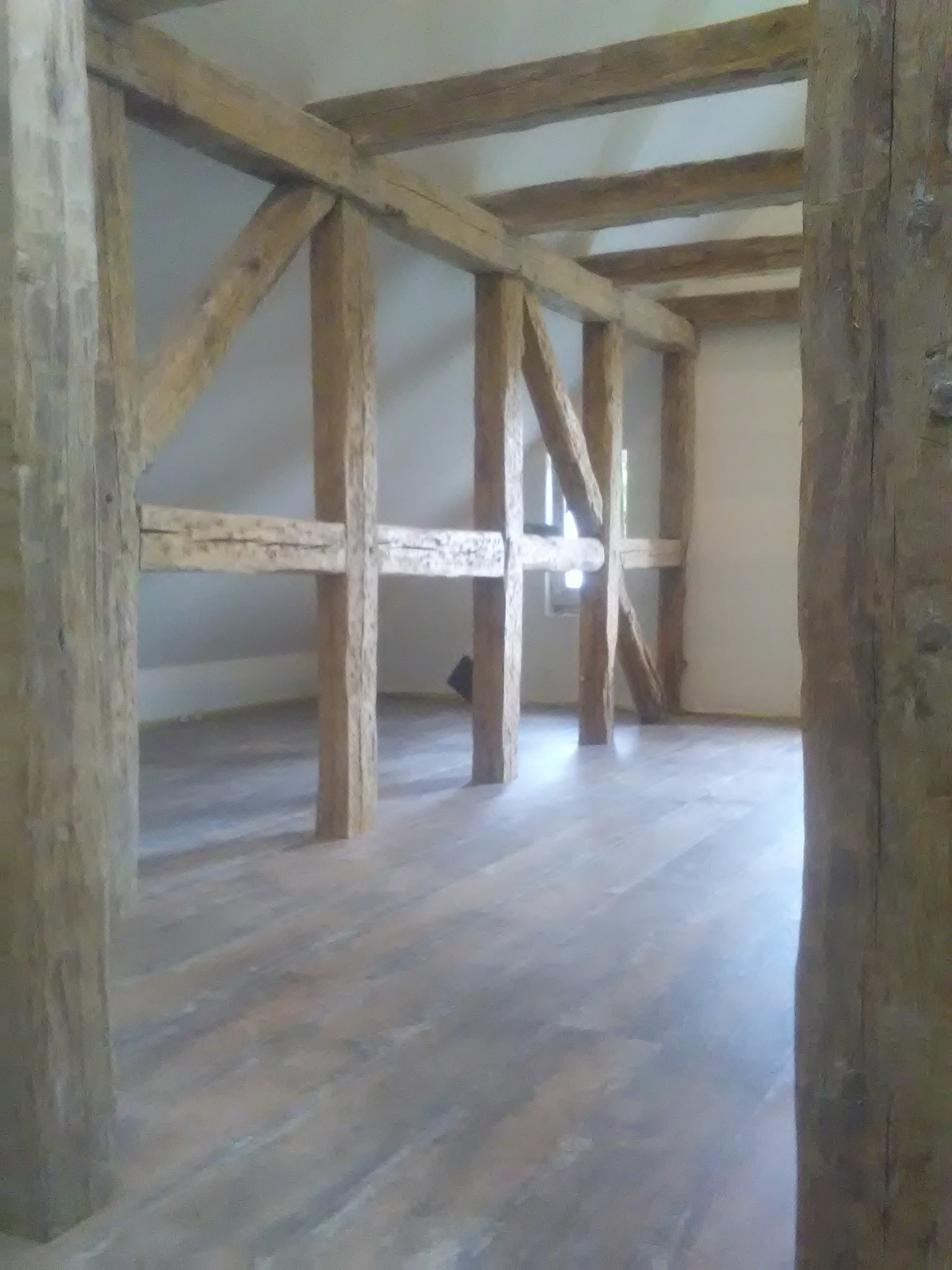 Surowa konstrukcja drewniana z widocznymi belkami nośnymi wewnątrz budynku, z drewnianą podłogą i oknem w tle.