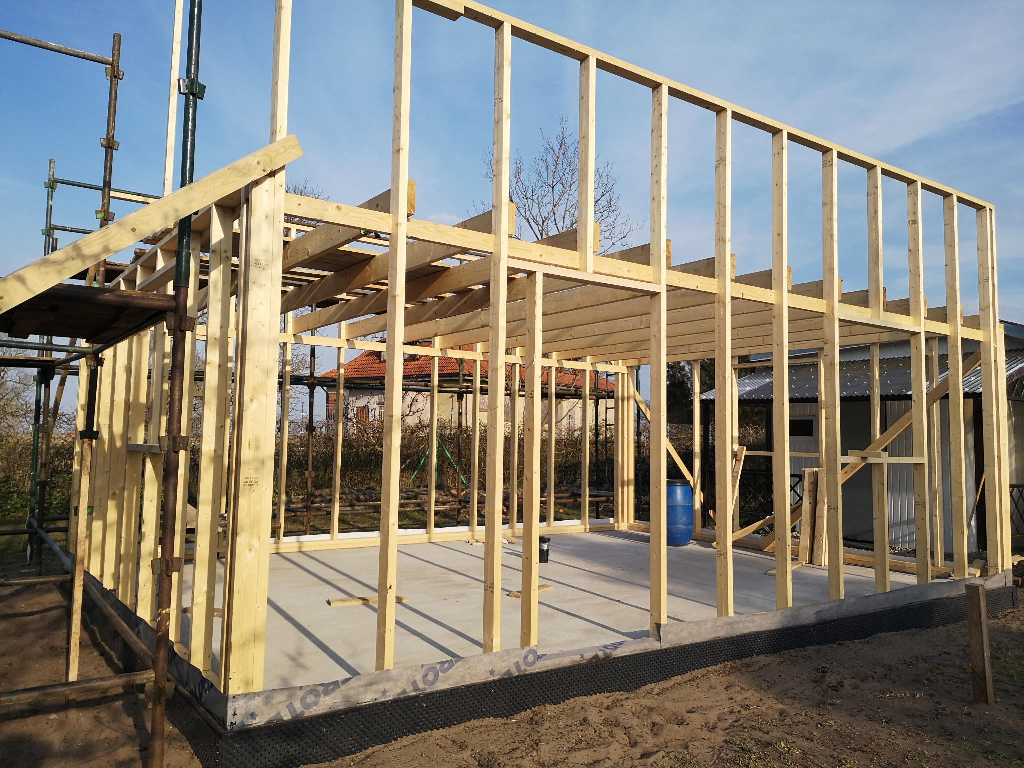 Wznoszenie konstrukcji szkieletowej domu z widocznym fundamentem, izolacją przeciwwilgociową i niebieską beczką w tle.