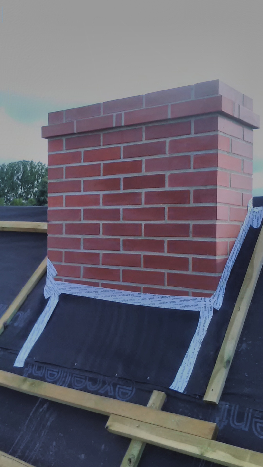 Murowany komin z cegły na dachu pokrytym czarną membraną, zabezpieczony taśmą uszczelniającą SIGA Wigluv, widoczne elementy drewnianej konstrukcji dachu.