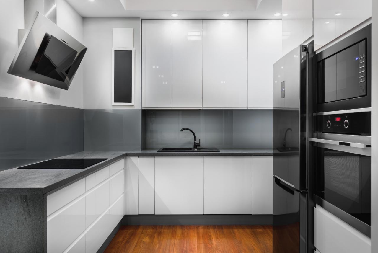 Nowoczesna kuchnia z białymi, lakierowanymi frontami, szarym blatem, drewnianą podłogą i zabudowanym sprzętem AGD.