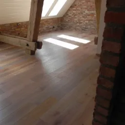 Jasne panele podłogowe ułożone w pokoju na poddaszu z drewnianymi belkami konstrukcyjnymi i ścianą z czerwonej cegły, oświetlone promieniami słońca wpadającymi przez okna dachowe.