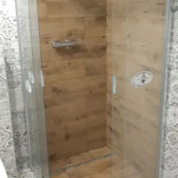 Kabina prysznicowa z drewnopodobnymi płytkami na ścianach i podłodze, otoczona wzorzystymi płytkami w odcieniach szarości i bieli. Na szybie kabiny naklejka ostrzegawcza.