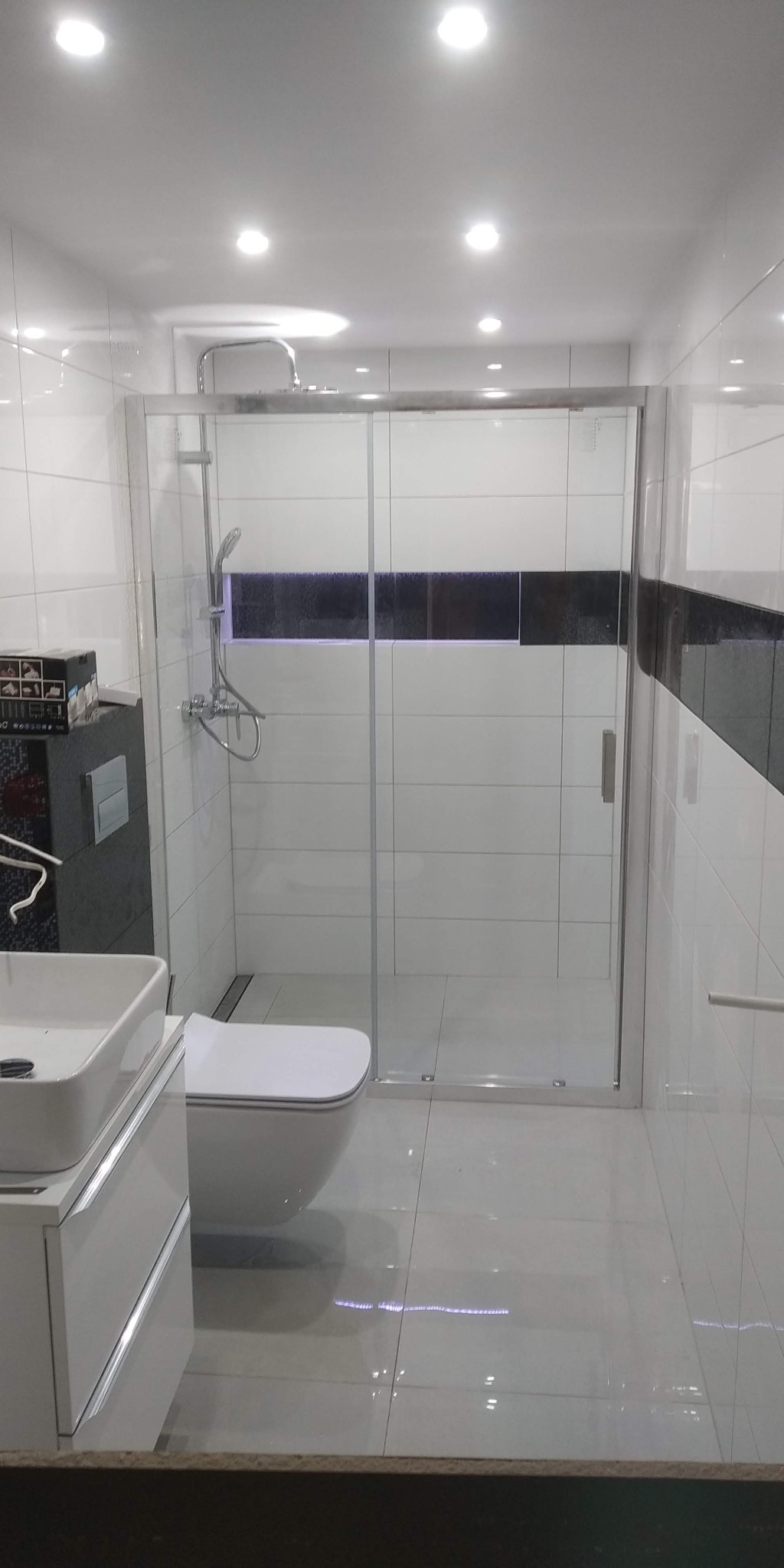 Nowoczesna łazienka z kabiną prysznicową z przesuwnymi drzwiami, wiszącą toaletą i umywalką nablatową na szafce, wykończona białymi płytkami z czarnym akcentem.