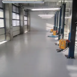 Jasnoszara posadzka żywiczna w hali warsztatowej z bramami segmentowymi i podnośnikami samochodowymi, oświetlona lampami jarzeniowymi.