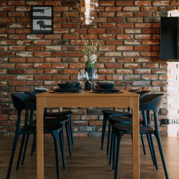Firma Patryk Gacek - Jadalnia z drewnianym stołem, czarnymi krzesłami i zastawą, na tle ściany z czerwonej cegły z nowoczesnym oświetleniem i minimalistycznym obrazem.