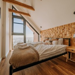 Firma Patryk Gacek - Jasna sypialnia na poddaszu z drewnianą ścianą dekoracyjną, widokiem na miasto i drewnianymi belkami stropowymi.