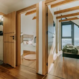 Widok przez otwarte, drewniane drzwi na wykończoną łazienkę z geometrycznymi płytkami i sypialnię na poddaszu z drewnianymi belkami stropowymi i dużym oknem.