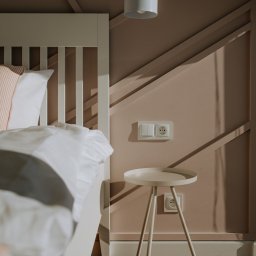 Firma Patryk Gacek - Jasna sypialnia z łóżkiem, stolikiem nocnym na trzech nogach i dekoracyjną ścianą w kolorze pudrowego różu z geometrycznymi listwami.