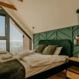 Sypialnia na poddaszu z dużym oknem z widokiem na miasto, zielona ściana z geometrycznym wzorem, łóżko z zielonym zagłówkiem i poduszkami, oraz wisząca lampa w stylu industrialnym.