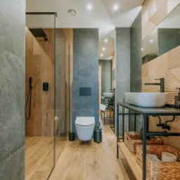 Nowoczesna łazienka z kabiną prysznicową typu walk-in, umywalką nablatową na metalowym stelażu, drewnopodobną podłogą i geometrycznymi płytkami na ścianie za lustrem.