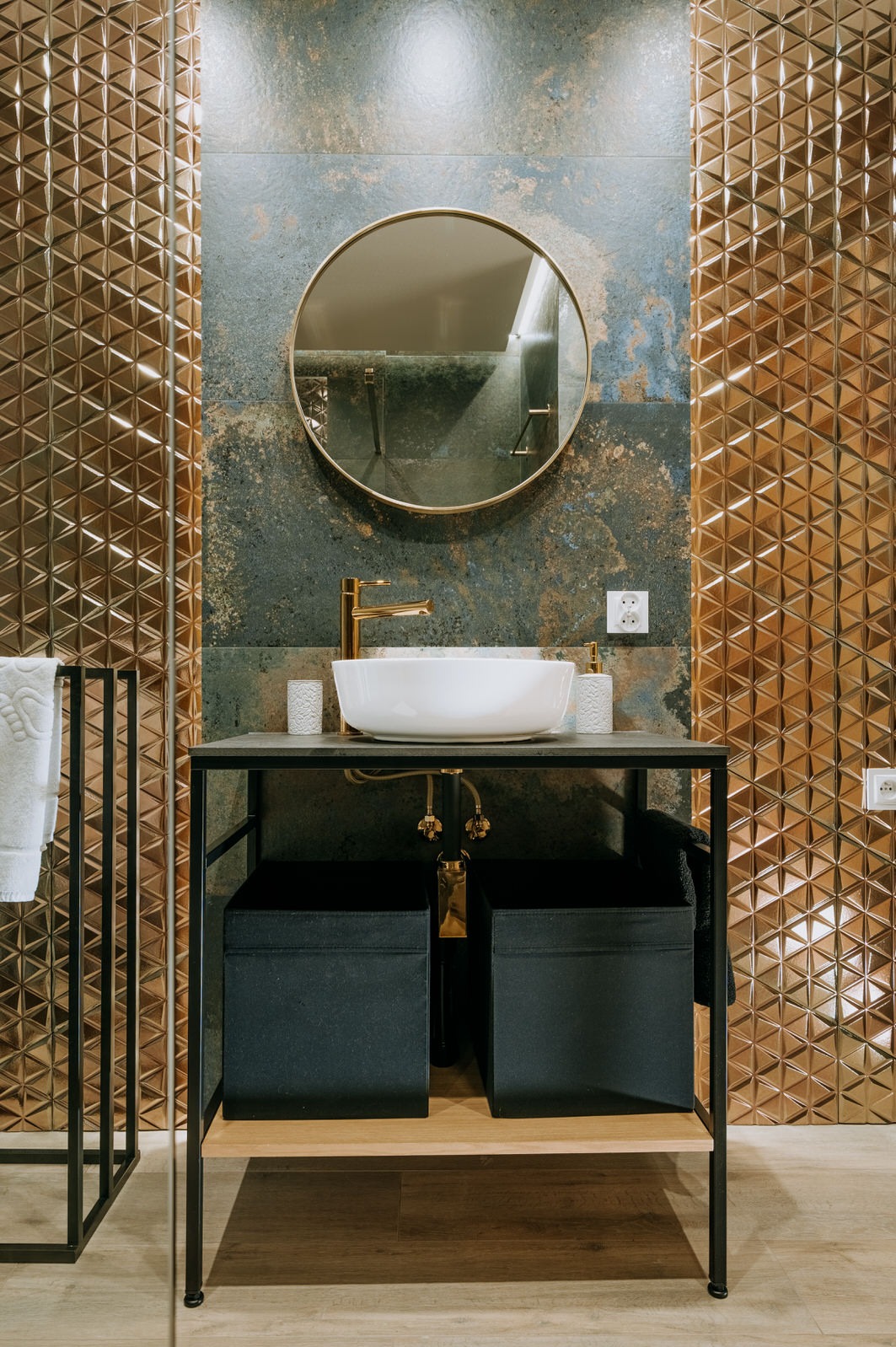Nowoczesna łazienka z umywalką nablatową, złotą armaturą i geometrycznymi, miedzianymi płytkami na ścianie, uzupełniona okrągłym lustrem z cienką ramą.
