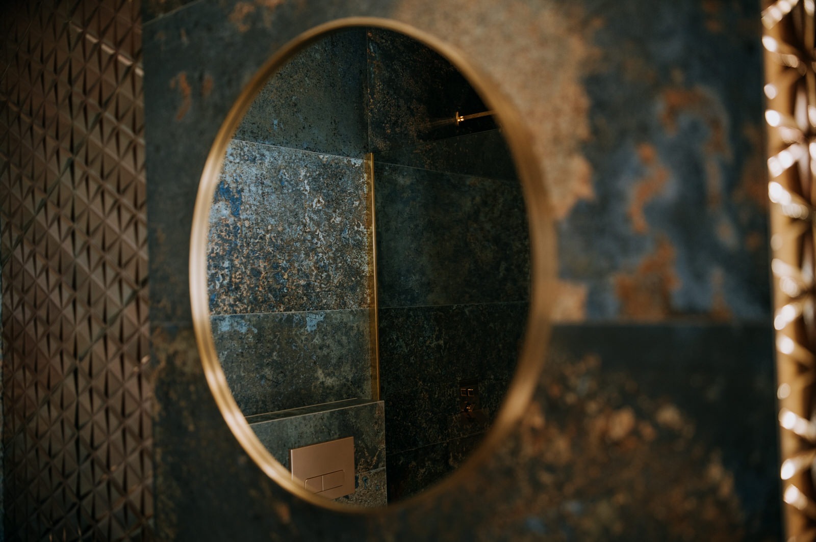 Okrągłe lustro z mosiężną ramą, odbijające fragment ściany wyłożonej płytkami imitującymi kamień w odcieniach szarości i niebieskiego z mosiężnymi akcentami, na ścianie po lewej stronie widoczna...
