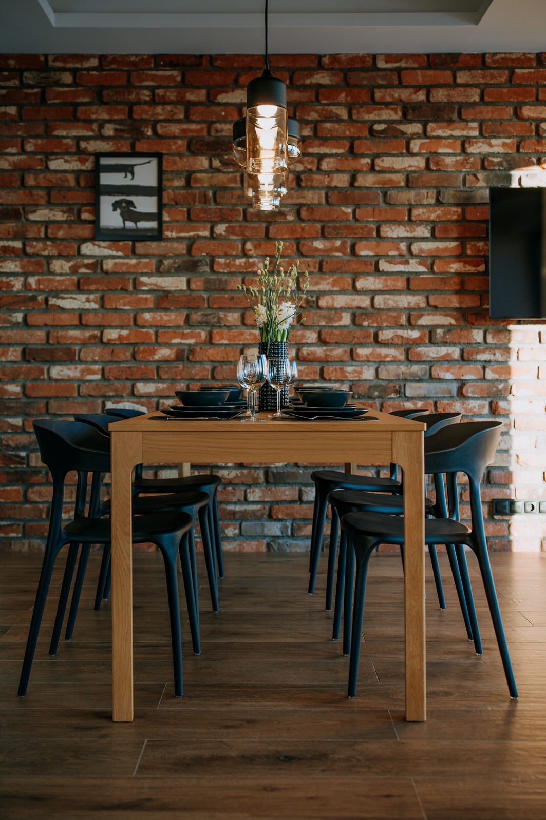 Jadalnia z drewnianym stołem, czarnymi krzesłami i zastawą, na tle ściany z czerwonej cegły z nowoczesnym oświetleniem i minimalistycznym obrazem.