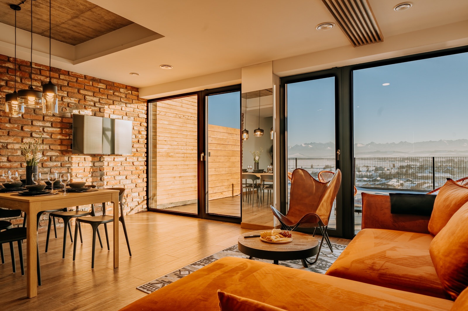 Jasne wnętrze apartamentu z ceglaną ścianą, drewnianym stołem jadalnym i widokiem na góry przez panoramiczne okna.