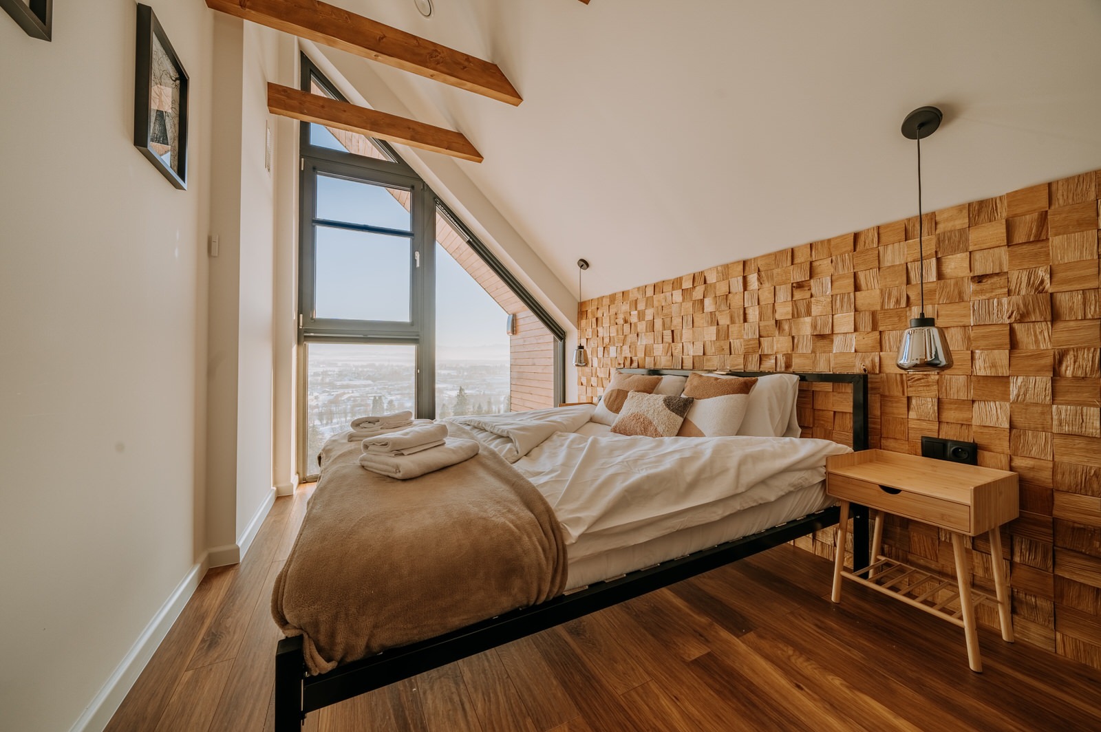 Jasna sypialnia na poddaszu z drewnianą ścianą dekoracyjną, widokiem na miasto i drewnianymi belkami stropowymi.