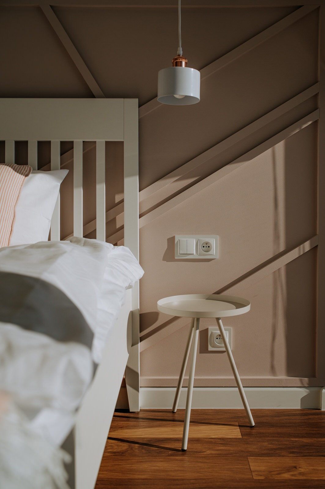 Jasna sypialnia z łóżkiem, stolikiem nocnym na trzech nogach i dekoracyjną ścianą w kolorze pudrowego różu z geometrycznymi listwami.