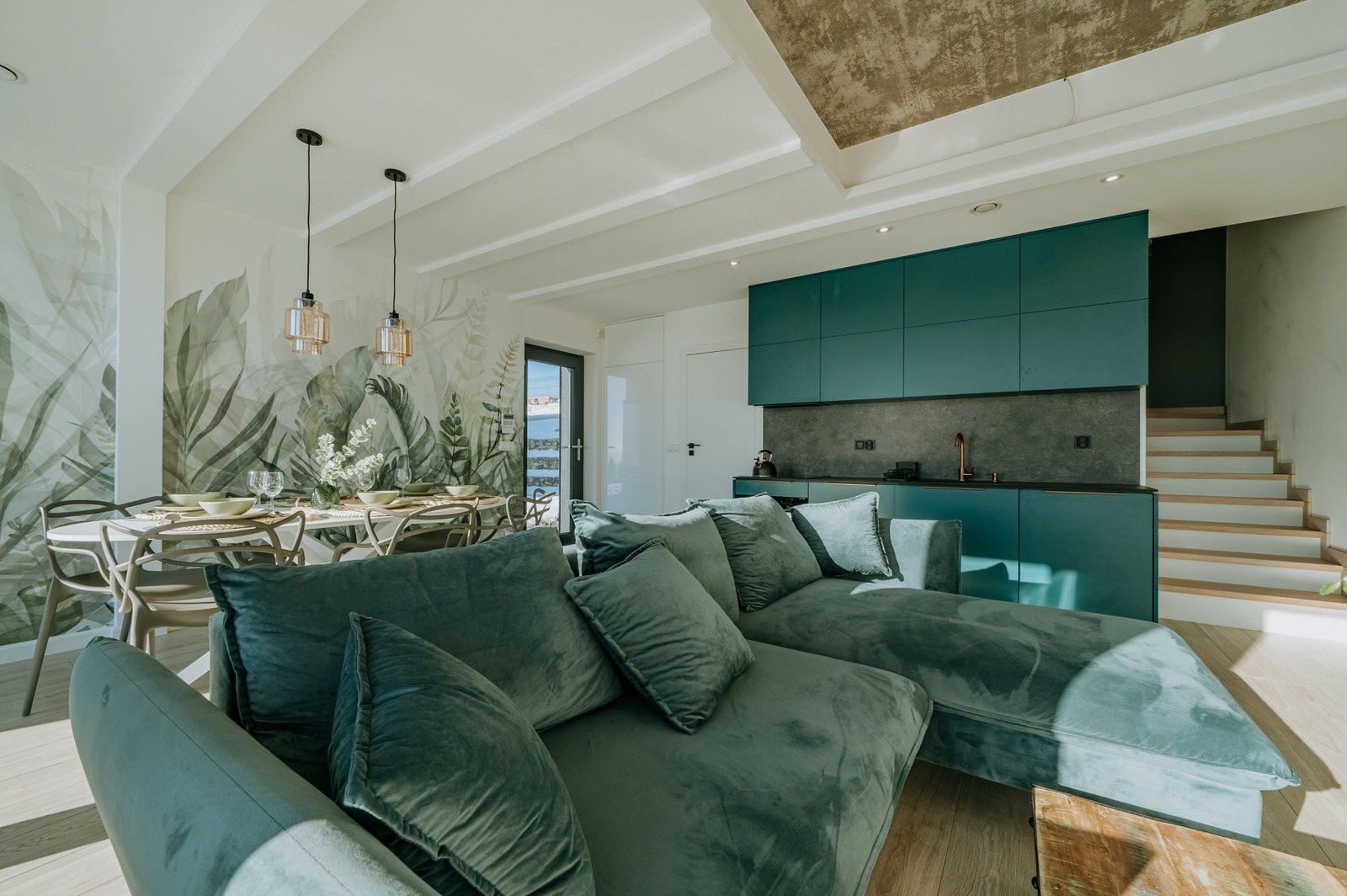 Nowoczesne wnętrze mieszkania z zieloną kuchnią, tapetą w liście i welurową sofą w odcieniach zieleni, oświetlone wiszącymi lampami nad stołem.