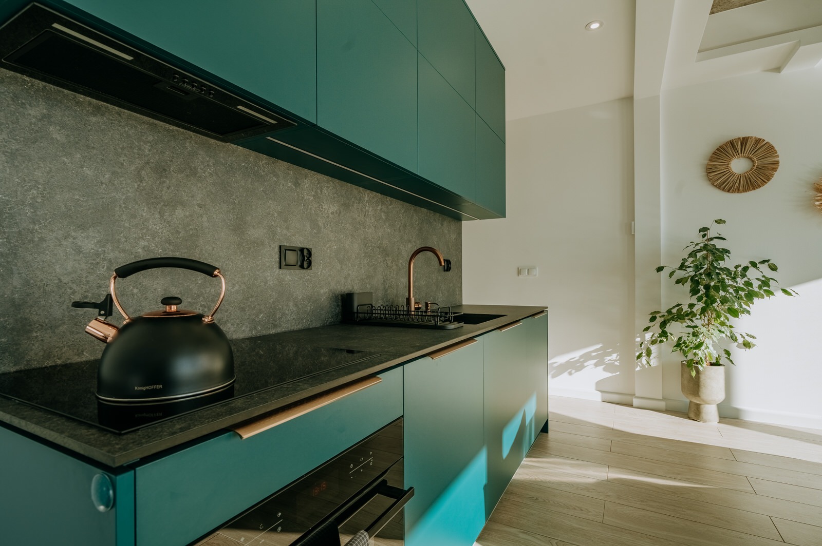 Nowoczesna kuchnia z turkusowymi szafkami, blatem z ciemnego kamienia i czajnikiem KönigHOFFER z miedzianymi akcentami, oświetlona promieniami słonecznymi padającymi na drewnianą podłogę.