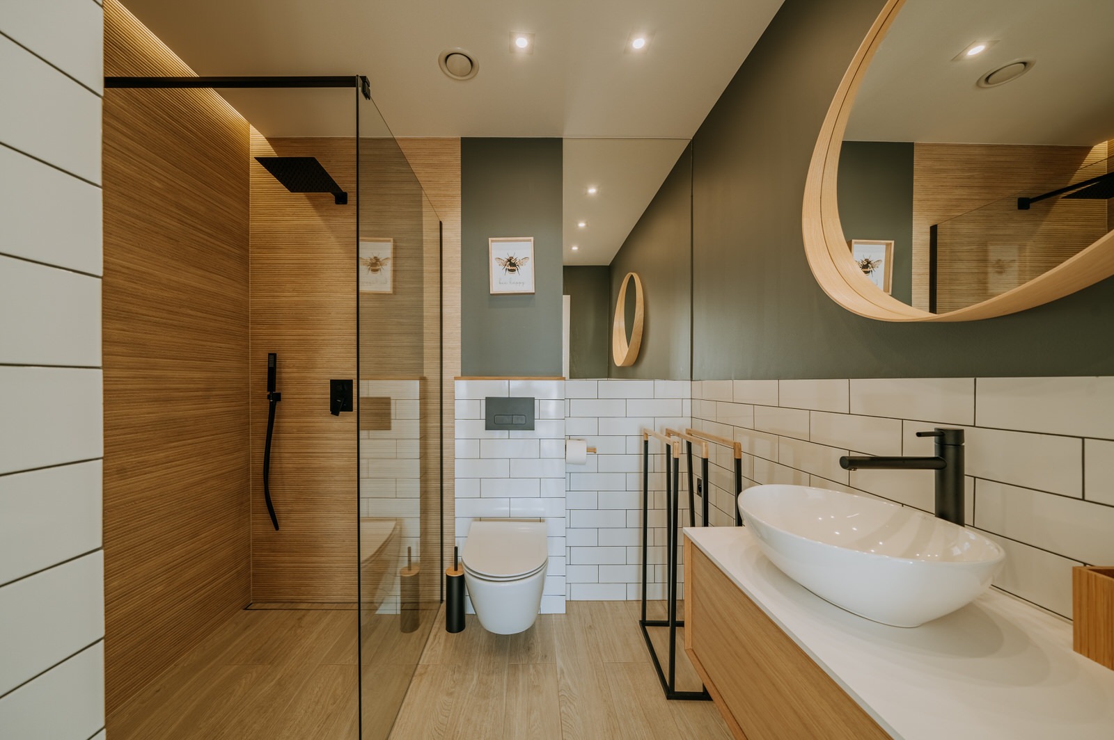 Nowoczesna łazienka z kabiną prysznicową typu walk-in, podwieszaną toaletą, umywalką nablatową w kształcie łzy i okrągłym lustrem z drewnianą ramą, zaakcentowana panelami imitującymi drewno...