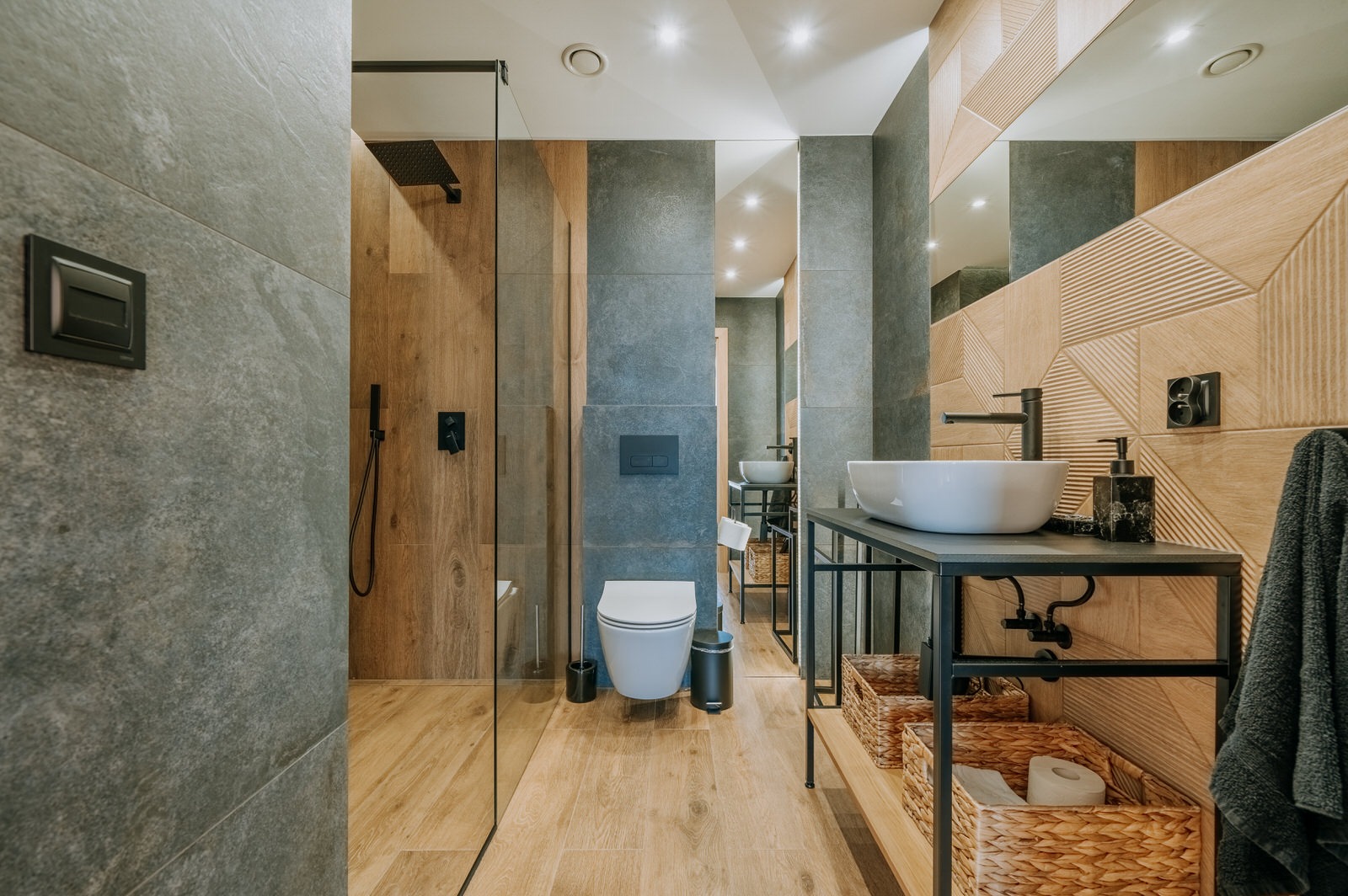 Nowoczesna łazienka z kabiną prysznicową typu walk-in, umywalką nablatową na metalowym stelażu, drewnopodobną podłogą i geometrycznymi płytkami na ścianie za lustrem.