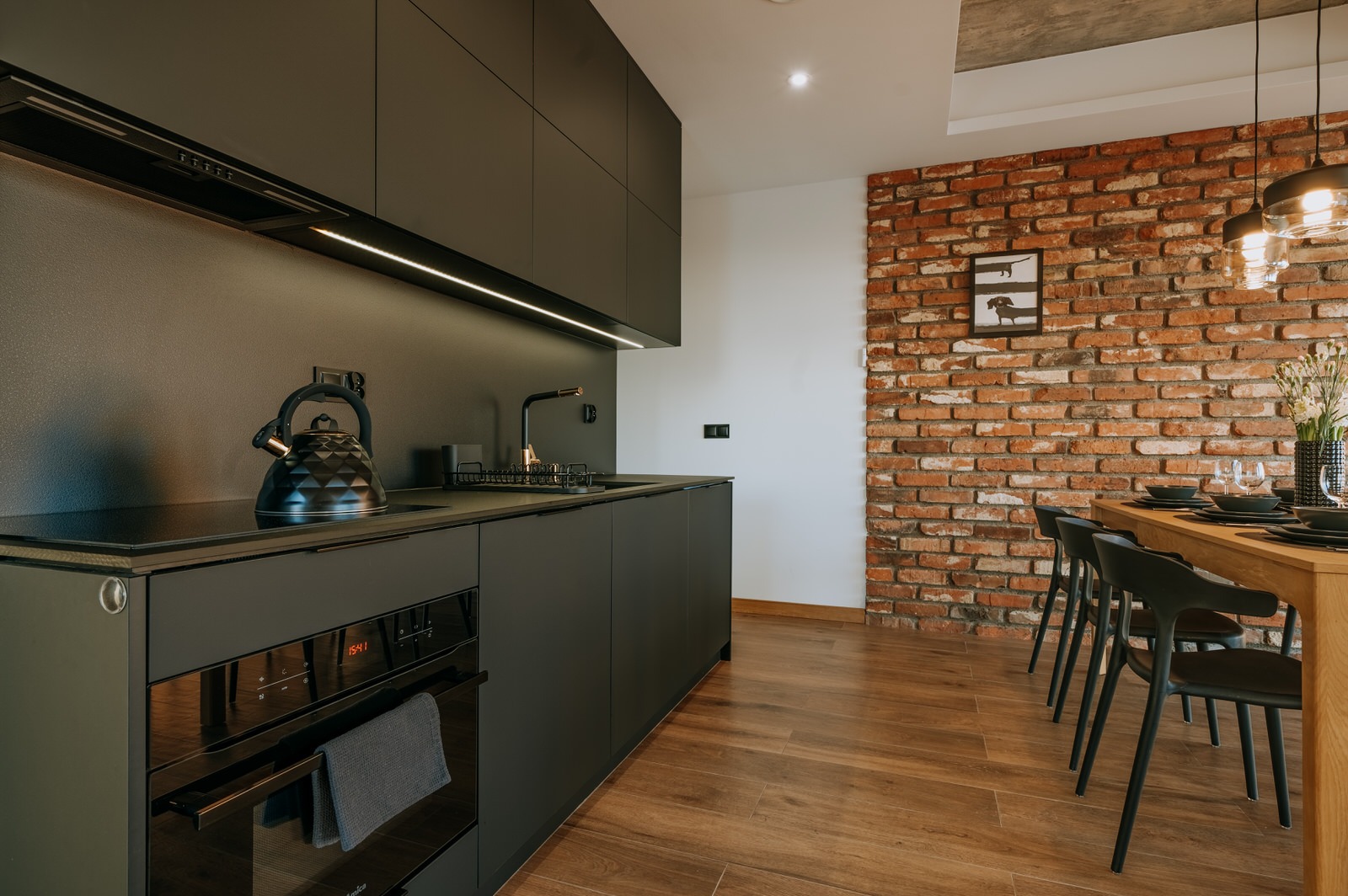 Nowoczesna kuchnia w ciemnej kolorystyce z ceglaną ścianą w tle, drewniane podłogi, czajnik na blacie i oświetlenie LED pod szafkami.