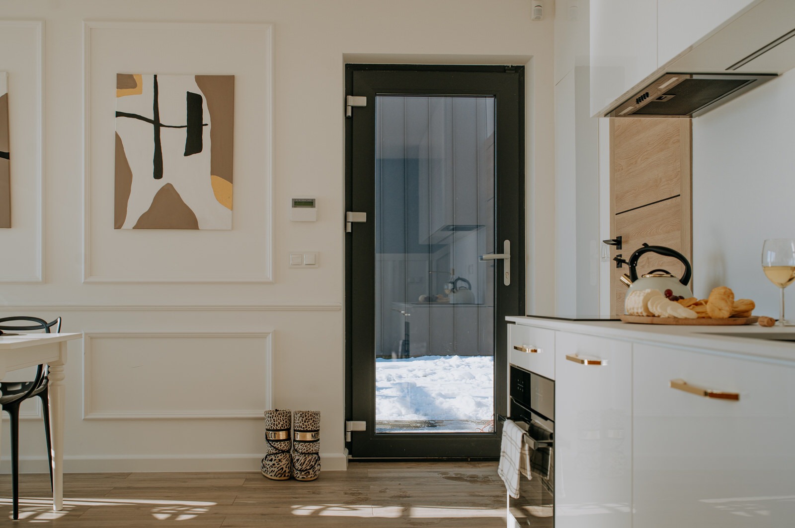 Jasne, nowocześnie urządzone wnętrze z białymi ścianami z dekoracyjnymi listwami, drewnianą podłogą, czarną ramą szklanych drzwi wejściowych, śniegiem na zewnątrz, abstrakcyjnymi obrazami...