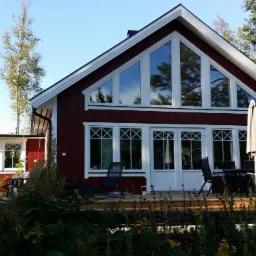 Czerwony dom jednorodzinny z białymi ramami okiennymi i drzwiami, z widocznymi oknami trójkątnymi na poddaszu i oknami z krzyżowymi szprosami na parterze, otoczony zielenią i drzewami, z lampą...