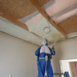 Pracownik w kombinezonie ochronnym aplikuje piankę PUR na drewnianą konstrukcję dachu, widoczne zabezpieczone folią fragmenty, przewody elektryczne i świeżo nałożona izolacja.