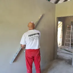 Mężczyzna w czerwonych spodniach i koszulce z logo firmy wygładza świeżo nałożony tynk na ścianie w surowym wnętrzu, widoczna drabina w tle.