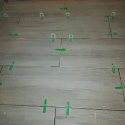 Układanie podłogi z płytek imitujących drewno, z użyciem zielonych klinów i białych poziomujących klipsów dystansowych, w trakcie prac wykończeniowych.