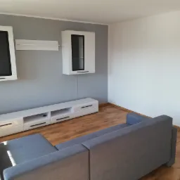 Nowoczesny salon z szarą ścianą, białymi wiszącymi szafkami z czarnymi ramkami, białą szafką RTV z czarnymi uchwytami i szarą sofą modułową na drewnianej podłodze.