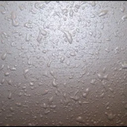 Szaro-brązowa, nierówna powierzchnia z licznymi, małymi, zastygłymi kroplami farby, tworzącymi chropowatą teksturę.