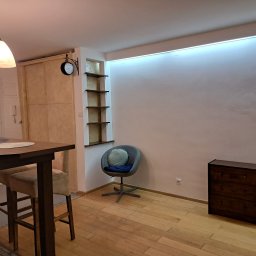 Firma Remontowo-Usługowa TATUŚ Bernard Rusin - Jasne wnętrze mieszkania z drewnianą podłogą, stołem barowym z krzesłami, szarą tapicerowaną pufą, wnęką z półkami i komodą w rogu, oświetlone listwą LED pod sufitem.