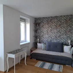 Pokój dzienny z tapetą w kwiaty, drewnianymi półkami, szarą sofą, białym biurkiem i drewnianą podłogą.