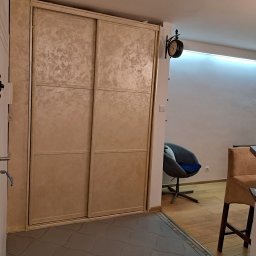 Firma Remontowo-Usługowa TATUŚ Bernard Rusin - Beżowa szafa przesuwna z dekoracyjną fakturą na frontach, widoczna w przedpokoju z płytkami podłogowymi w geometryczny wzór i fragmentem krzesła obrotowego w tle.