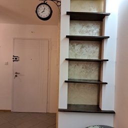 Firma Remontowo-Usługowa TATUŚ Bernard Rusin - Korytarz po remoncie: białe ściany, ciemne drewniane półki w zabudowie z dekoracyjną ścianą w odcieniach beżu i złota, drzwi wejściowe z zabezpieczeniami, zegar wiszący na ścianie.
