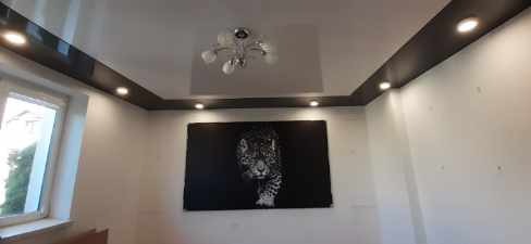 Błyszczący, biały sufit napinany z czarnymi listwami obwodowymi i wbudowanymi punktami świetlnymi, centralnie umieszczony żyrandol, na ścianie obraz z motywem pantery.