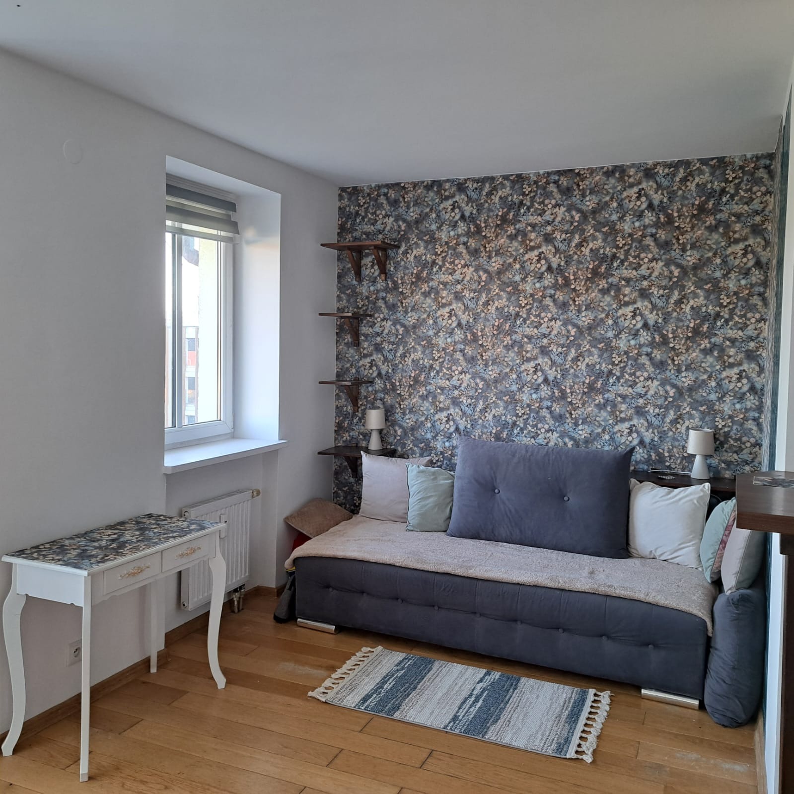 Pokój po remoncie z wzorzystą tapetą w odcieniach niebieskiego i brązowego, drewnianymi półkami, szarą sofą z poduszkami, dywanikiem w paski i białą toaletką z blatem pasującym do tapety.