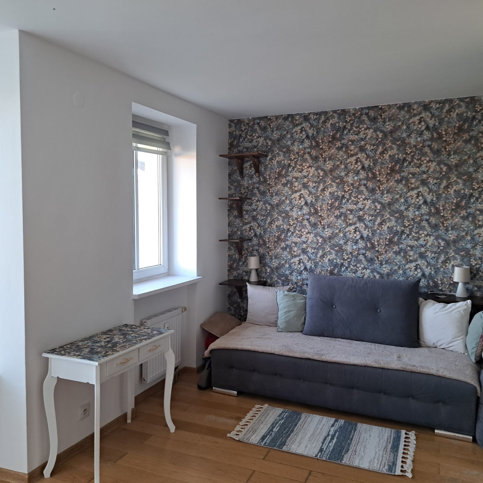 Pokój dzienny z tapetą w kwiaty, drewnianymi półkami, szarą sofą, białym biurkiem i drewnianą podłogą.