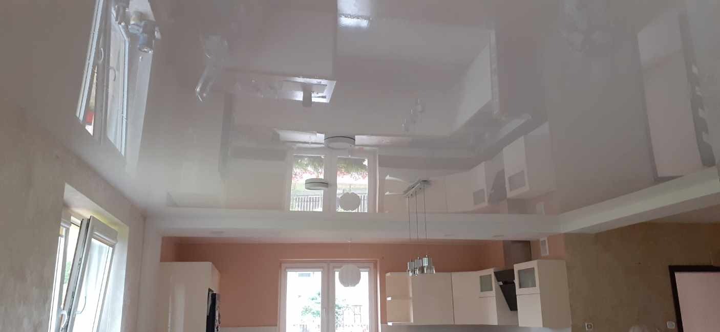 Błyszczący, biały sufit napinany odbijający światło z okien w nowoczesnej kuchni z białymi szafkami.