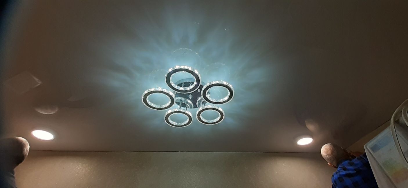 Zamontowany, nowoczesny żyrandol sufitowy z sześcioma okrągłymi elementami z kryształkami, oświetlający pomalowany sufit z dodatkowymi punktowymi lampami, widoczni dwaj instalatorzy podczas pracy.